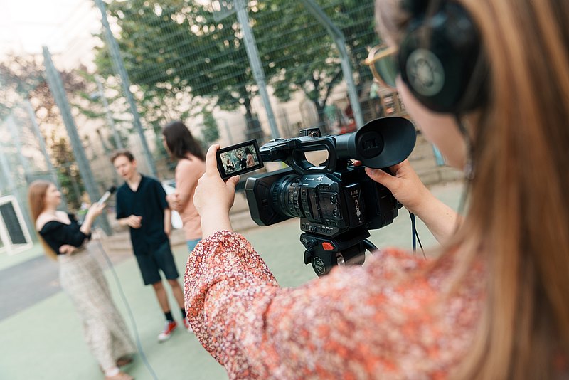 Eine junge Person mit langen Haaren filmt eine Interview-Szene. Die Szene ist durch den Bildschirm der Kamera nochmal sichtbar.