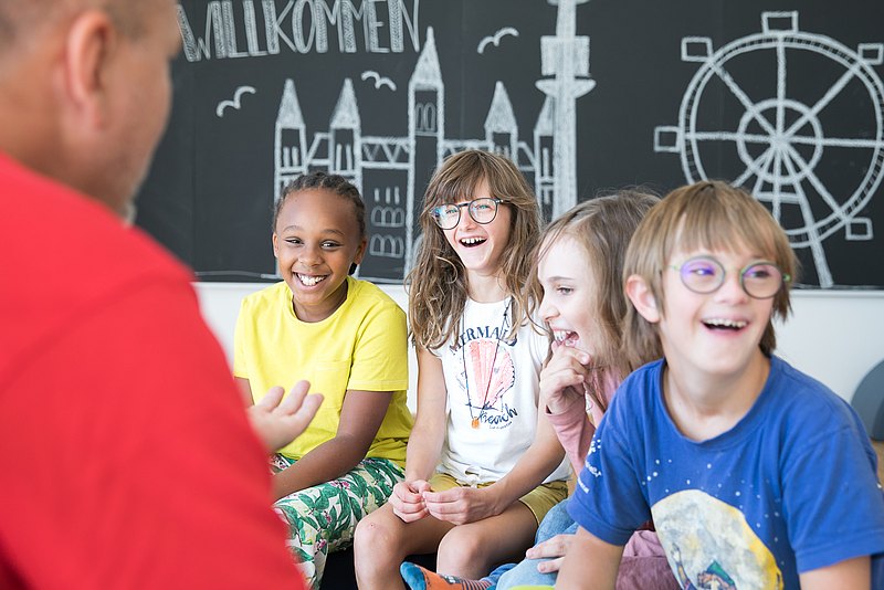 Lachende Kinder vor einer Tafel mit weißem Riesenrad. Mit den Kindern spricht eine erwachsene Person im roten Shirt. 