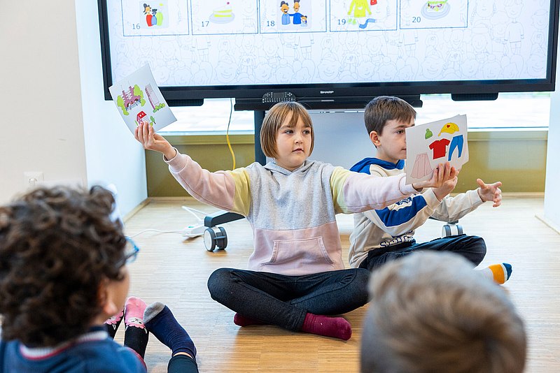 Am Boden sitzen Kinder, eins davon hat Bildtafeln in der Hand und hät sie mit weit ausgestreckten Händen