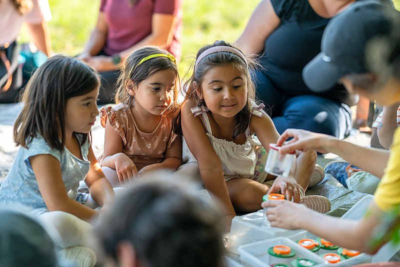 Kinder sitzen draußen im Gras und schauen interessiert auf einen Behälter, den ein anderes Kind in der Hand hat