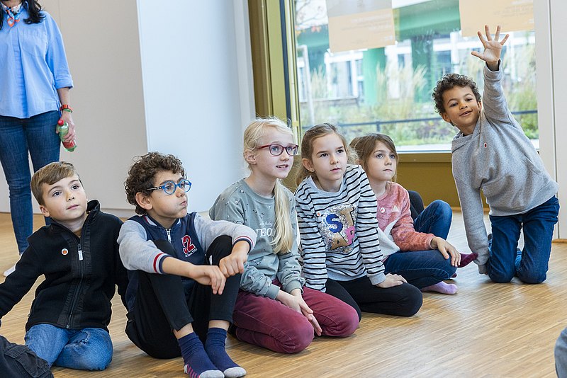 Kinder sitzen am Boden. Eins zeigt auf, so als ob´s unbedingt drankommen will
