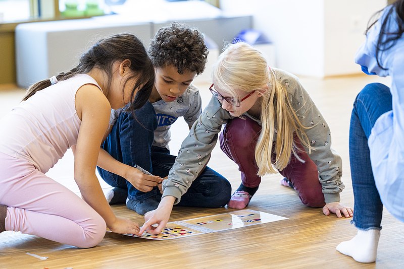 Drei Kinder sitzen am Boden vor einem Spiel und schauen und zeigen auf die Unterlage
